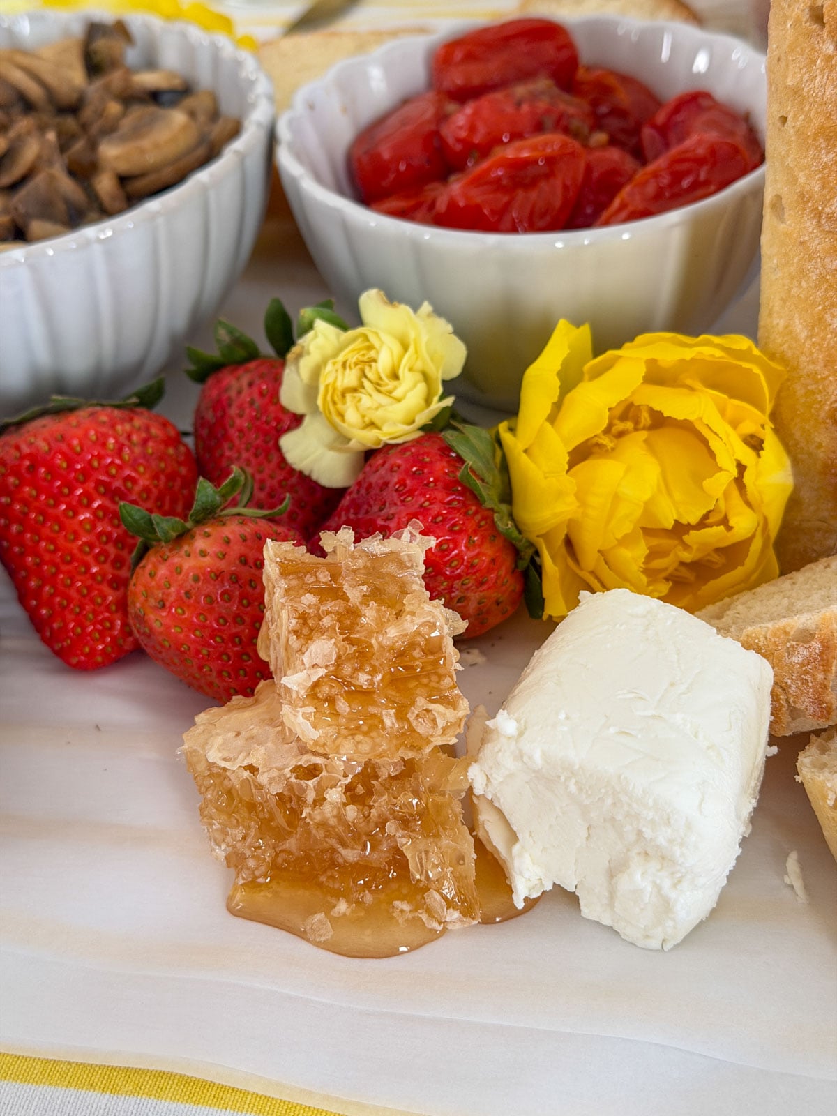 Compound butter toast party sweet side grazing board with honeycomb, cream cheese, fresh strawberries, yellow roses and tulips, roasted cherry tomatoes, sauteed mushrooms, and sliced baguette.