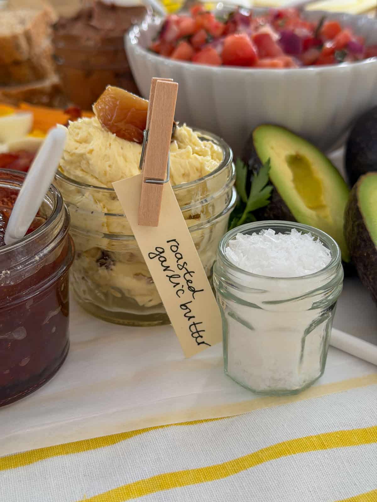 Small glass jar of roasted garlic compound butter with a handwritten clothespin label beside a jar of flaky sea salt on a white grazing surface with avocado and bruschetta topping in the background.