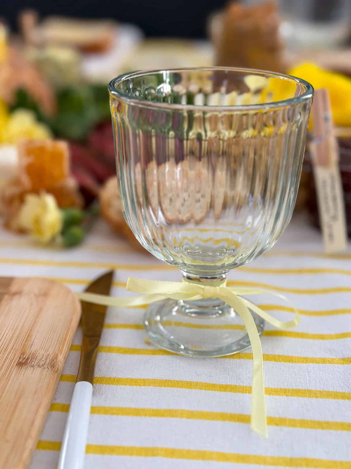 Clear ribbed goblet wine glass with a yellow satin ribbon bow tied at the stem on a yellow striped tablecloth with the compound butter grazing table in soft focus behind.