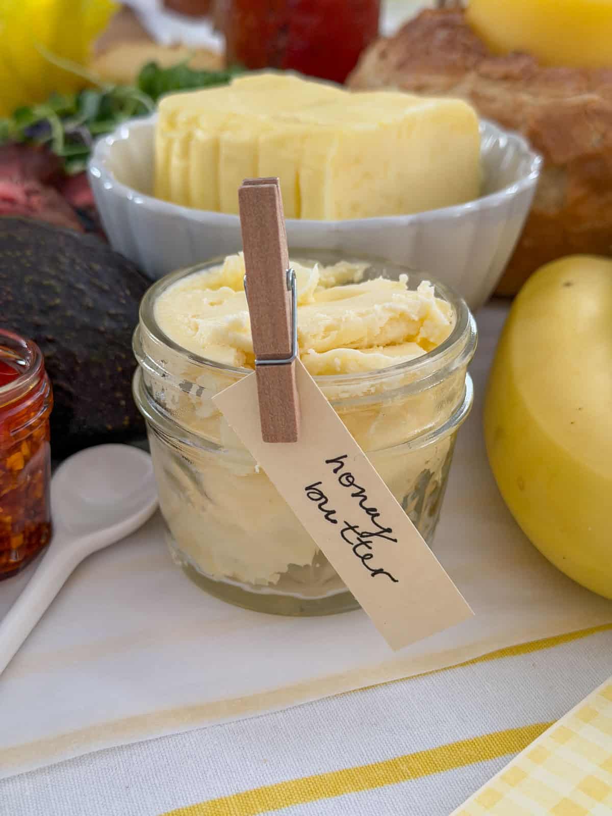 Jar of whipped honey butter with a clothespin label on a white parchment grazing surface with a block of sliced French butter in a white scalloped bowl behind.