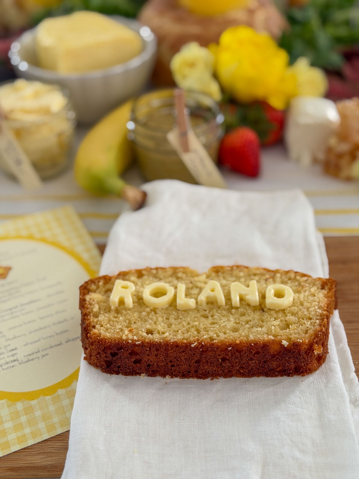 Slice of cornbread with butter letter tiles spelling ROLAND used as a name card place setting on a white linen napkin with house recommendations menu card, banana, and grazing board spread in background.