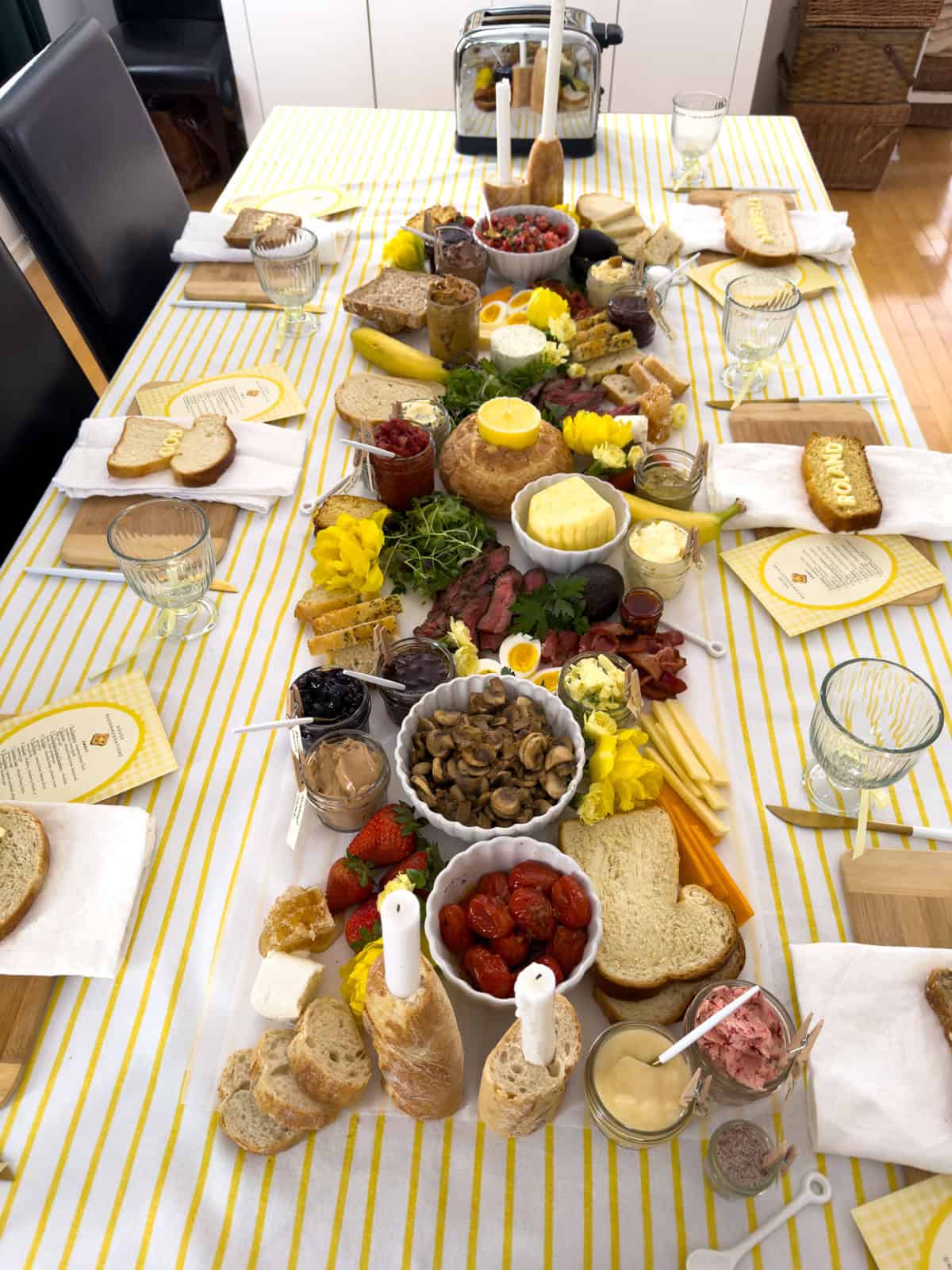 Overhead view of butter themed toast party dining table with yellow striped tablecloth showing full compound butter grazing spread, Roland cornbread name bread and Dannyelle sourdough name bread place settings, butter candle in sourdough boule centerpiece, baguette candle holders, and toaster at the far end.