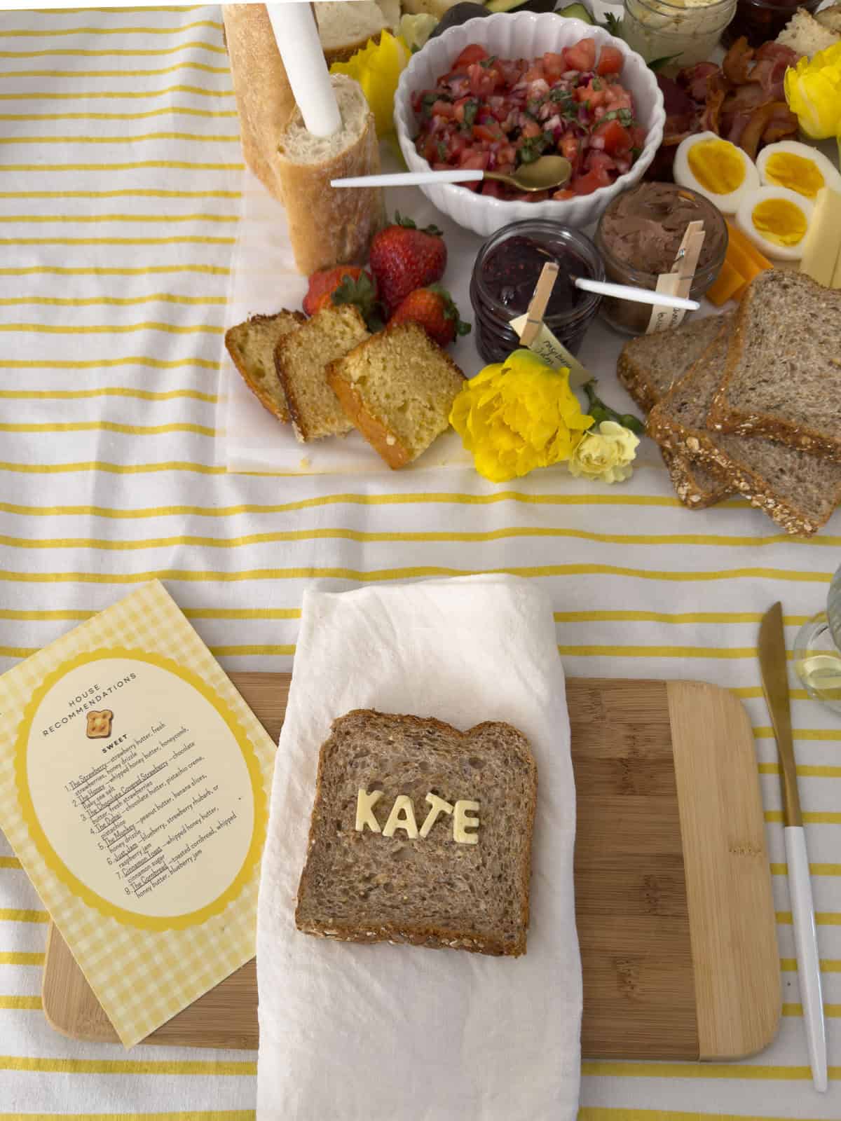 Kate butter letter name bread place setting with house recommendations menu card on bamboo cutting board beside grazing board with bruschetta topping bowl, strawberries, cornbread slices, and yellow flowers.