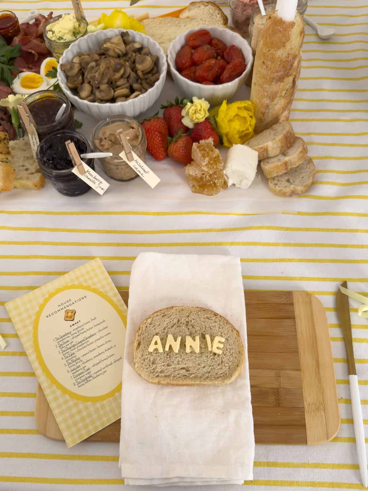 Overhead view of Annie butter letter name bread place setting on bamboo cutting board with house recommendations menu card, blueberry vanilla jam jar, hazelnut creme jar, roasted cherry tomatoes, fresh strawberries, honeycomb, sliced baguette, and yellow tulip visible on the grazing table behind.