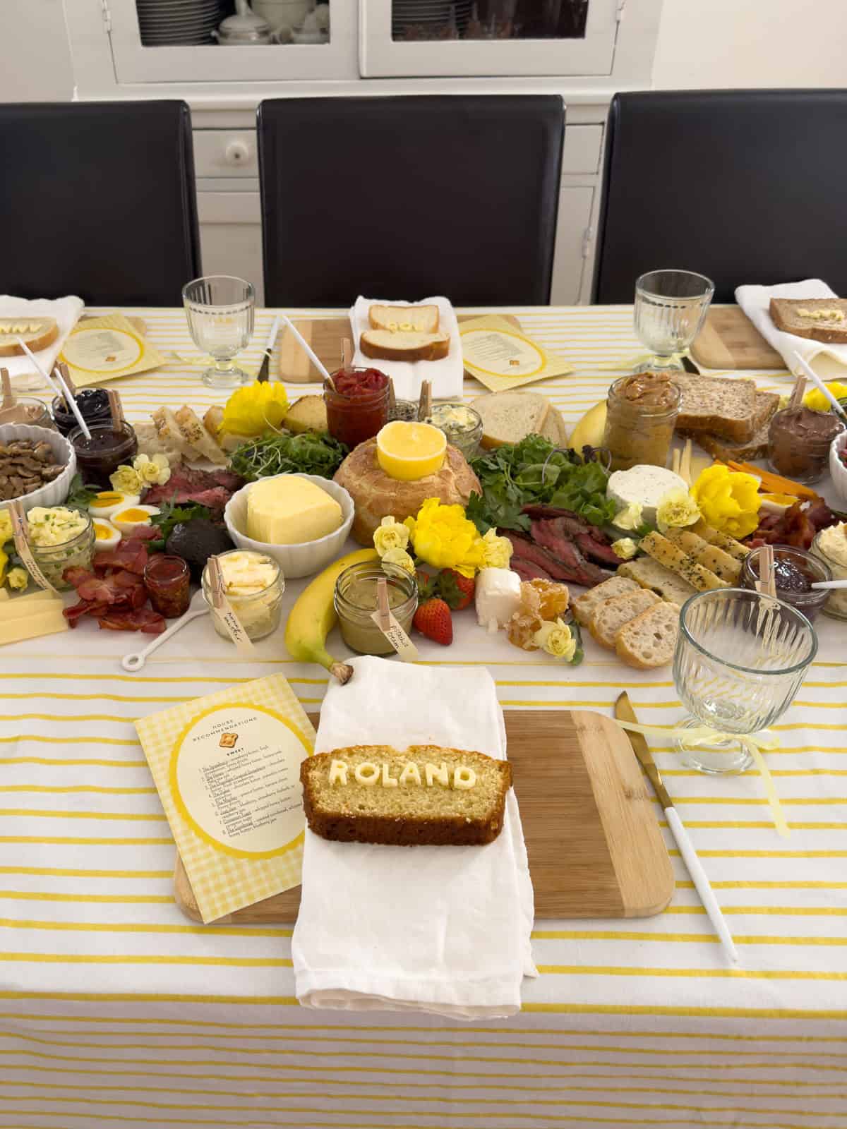 Butter toast dinner party table set for six with Roland cornbread name bread in foreground, full grazing spread with butter block, steak, flowers, and labeled jars stretching the length of the table.