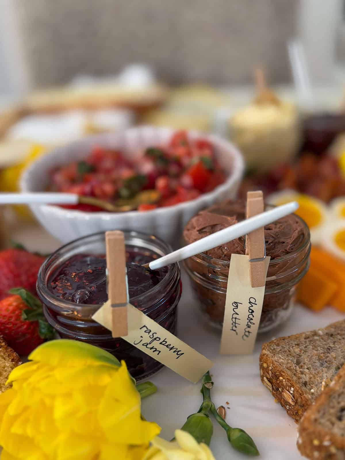 Small glass jar of chocolate compound butter with a handwritten clothespin label beside a jar of raspberry jam on a white parchment grazing surface with fresh strawberries, yellow tulip, sliced whole grain bread, bruschetta topping bowl, and soft boiled egg visible in background.