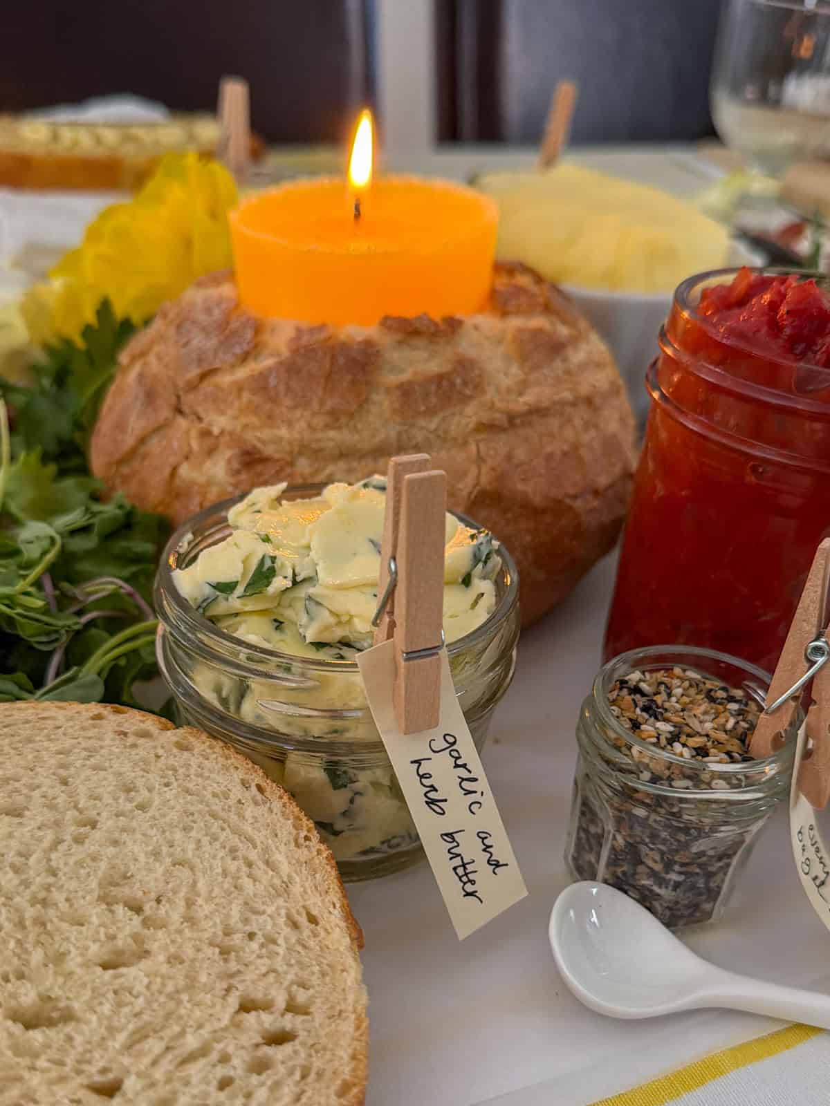 Jar of garlic herb compound butter with a handwritten clothespin label in the foreground with a burning butter candle in a sourdough boule, tomato jam jar, everything bagel seasoning jar, fresh mixed greens, and sliced sourdough bread visible around it on the grazing table.