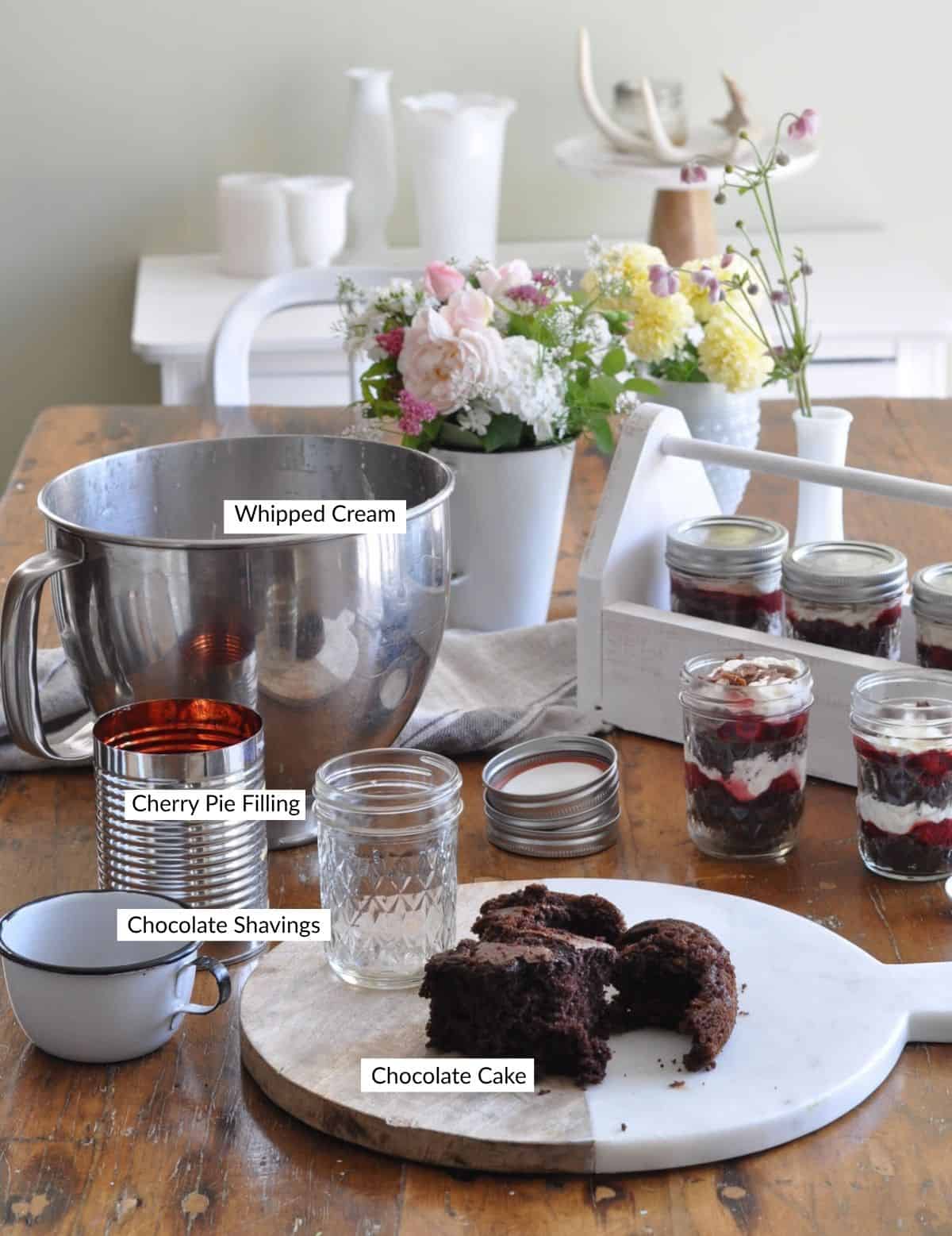 Black forest trifle ingredients, with mason jars in a white wooden carrier on a farmhouse table with stand mixer bowl, chocolate cake, and fresh flowers.