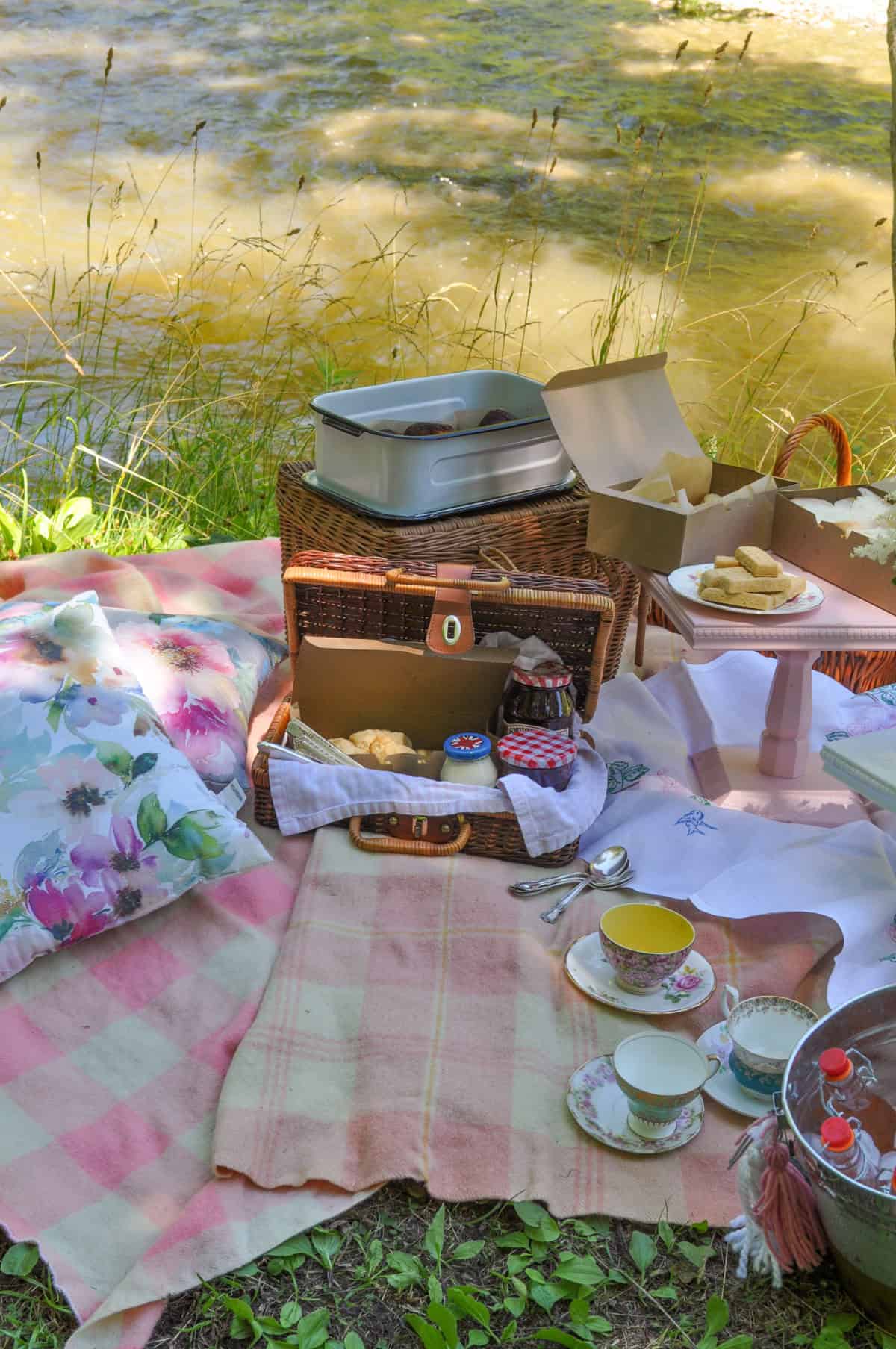 Tea party picnic spread with open basket, teacups, and lemonade by the river.