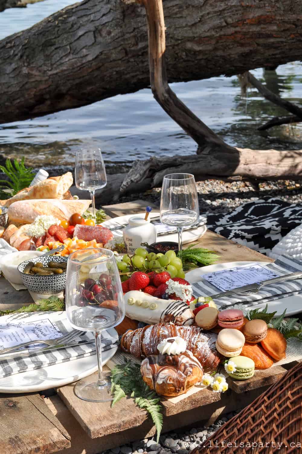 French inspired picnic charcuterie board on a wooden pallet table beside the lake with wine glasses, French pastries, fresh fruit, cured meats, and a fallen log and water in the background.