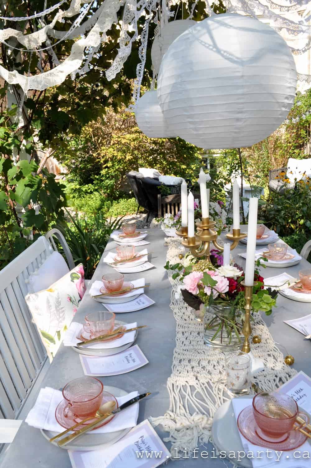White backyard pergola with climbing pink roses, lace ribbon swags, and hanging paper lanterns framing a garden tea party setting at dusk.
