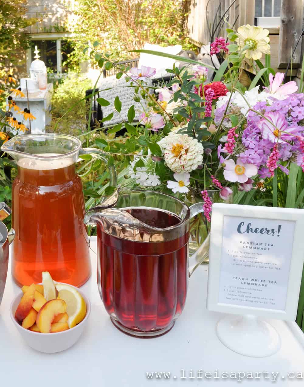 Garden tea party iced tea mocktail station with pitchers of passion tea lemonade and peach white tea lemonade, fresh peach and lemon garnishes, printed recipe card, and large fresh garden flower arrangement.