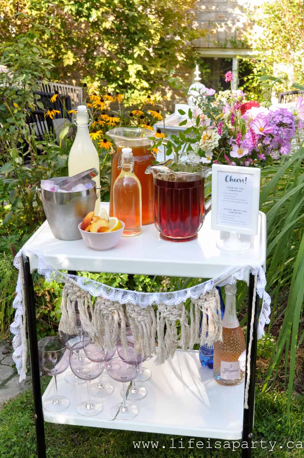 Garden tea party iced tea bar cart with pitchers of passion tea lemonade and peach white tea lemonade, silver ice bucket, purple wine glasses, and fresh garden flower arrangement in a sunny backyard setting.
