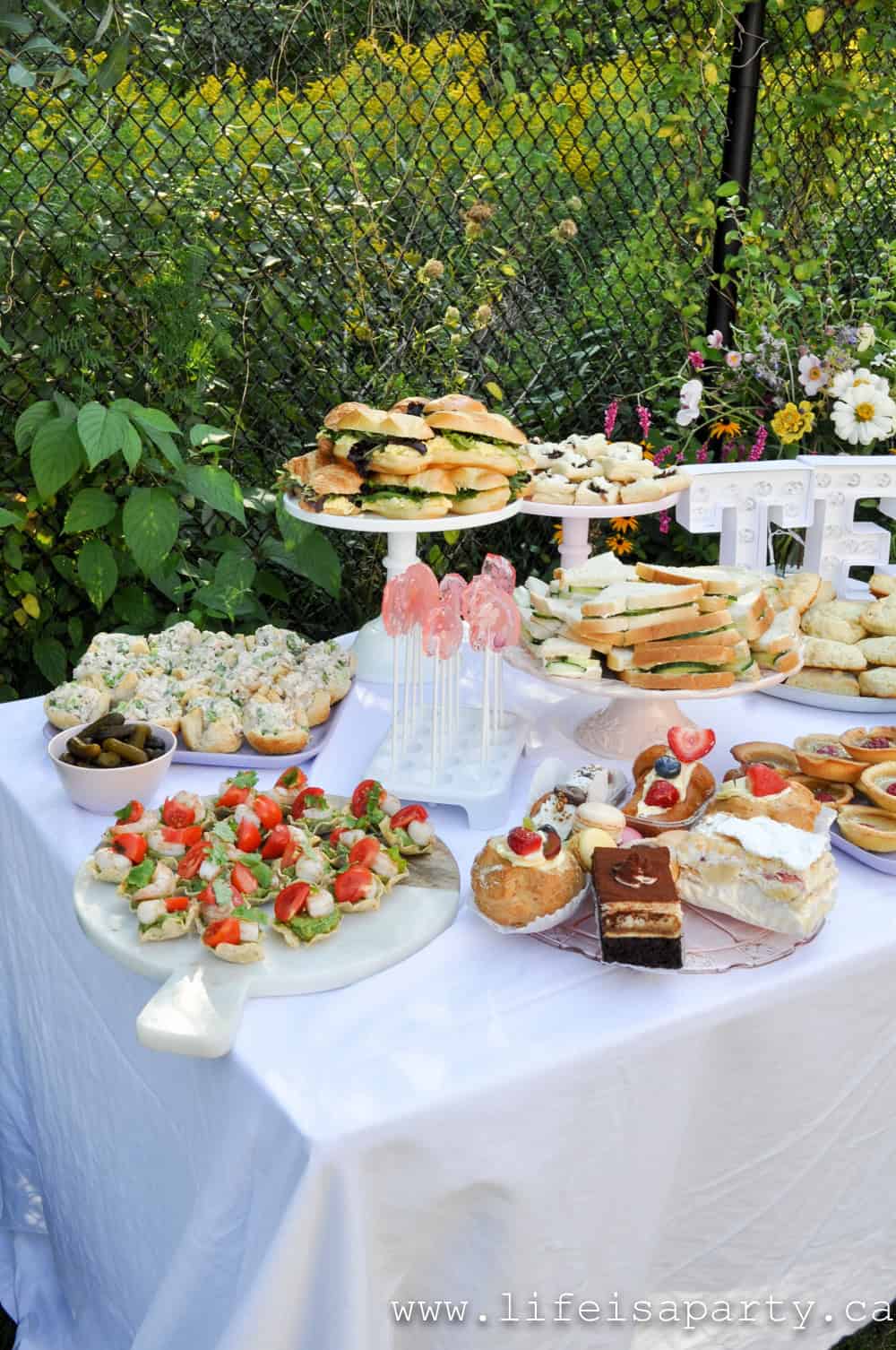 Garden tea party food table with cucumber sandwiches on a tiered cake stand, chicken salad cups, avocado shrimp tostadas, floral lollipops, raspberry tarts, and assorted sweets on a white tablecloth with garden backdrop.