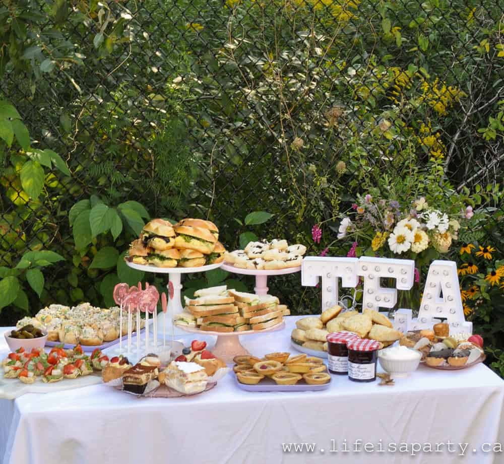 Garden tea party food table with tiered stands of finger sandwiches, chicken salad cups, avocado tostadas, floral lollipops, scones with jam and cream, raspberry tarts, and French pastries beside illuminated marquee TEA letters and fresh wildflower arrangement.