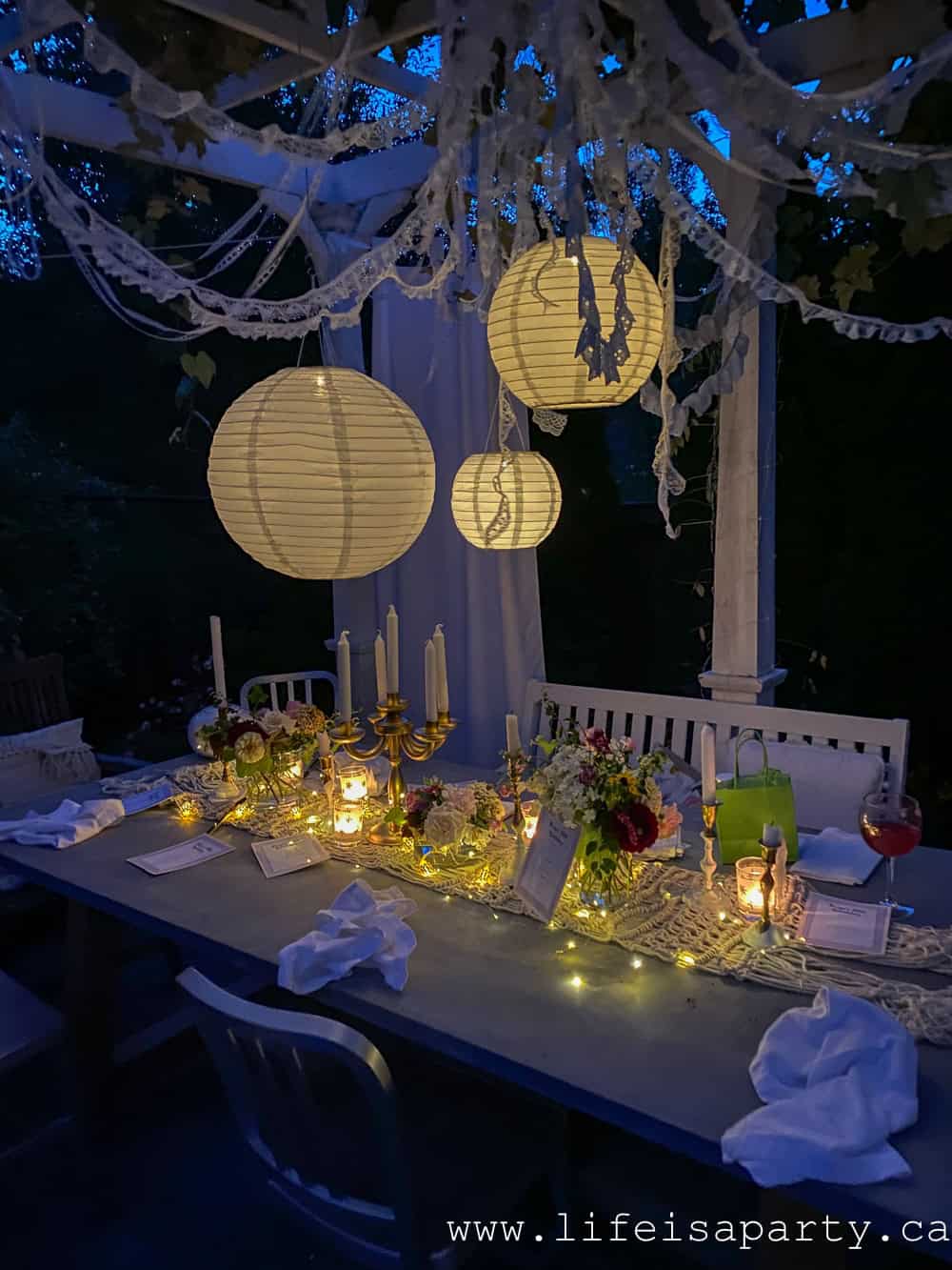 Garden tea party table glowing at night under lit paper lanterns with fairy lights along the macrame runner, gold candelabra with candles, and fresh flowers under a white pergola.