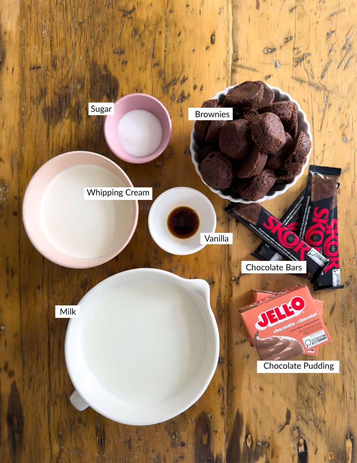 Labeled ingredients for mini chocolate brownie trifles including two-bite brownies, Jello instant chocolate pudding, Skor bars, whipping cream, milk, sugar, and vanilla arranged on a wood table.