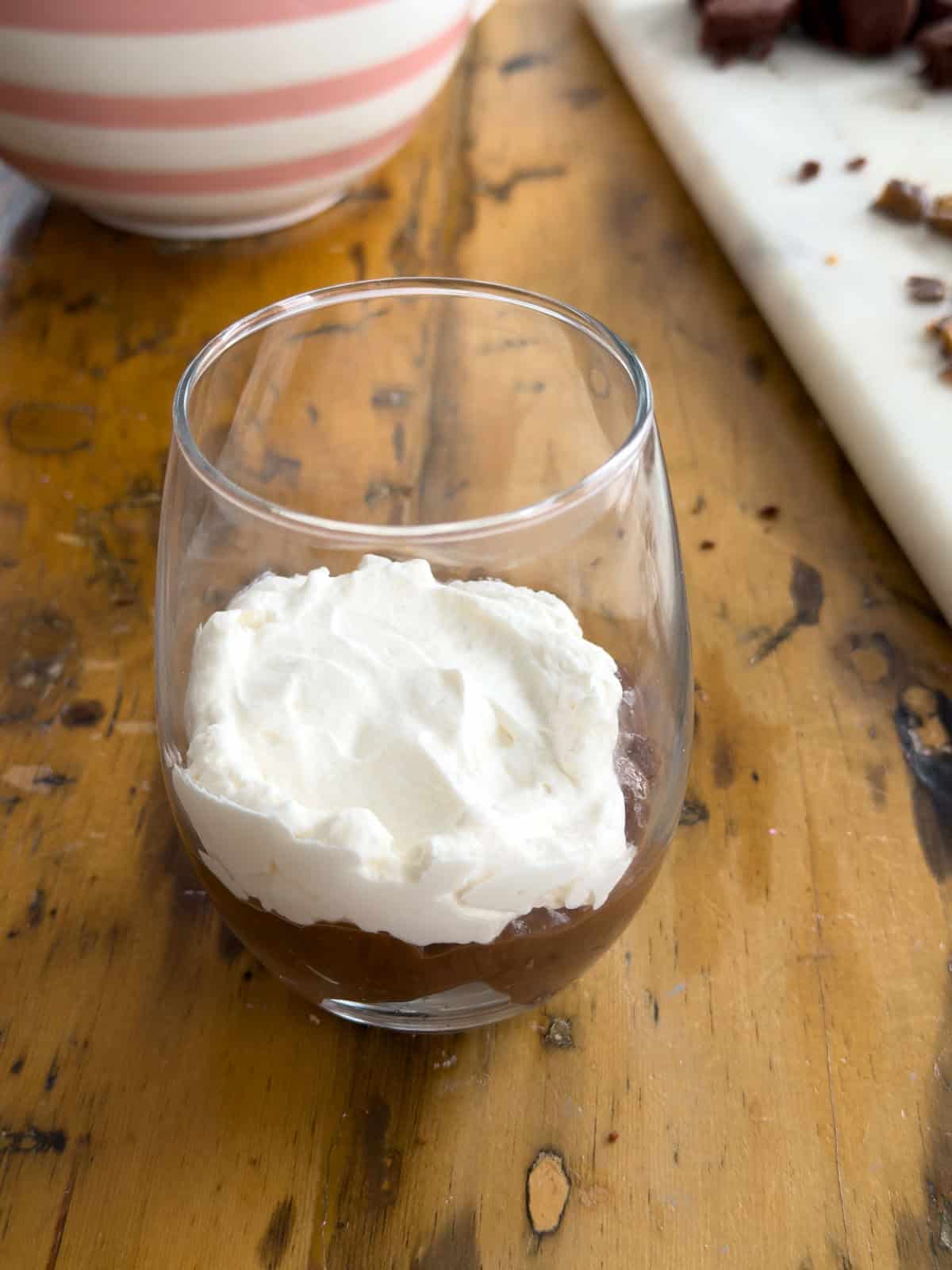 Homemade whipped cream spread over the chocolate pudding layer in a clear glass for mini chocolate brownie trifles.
