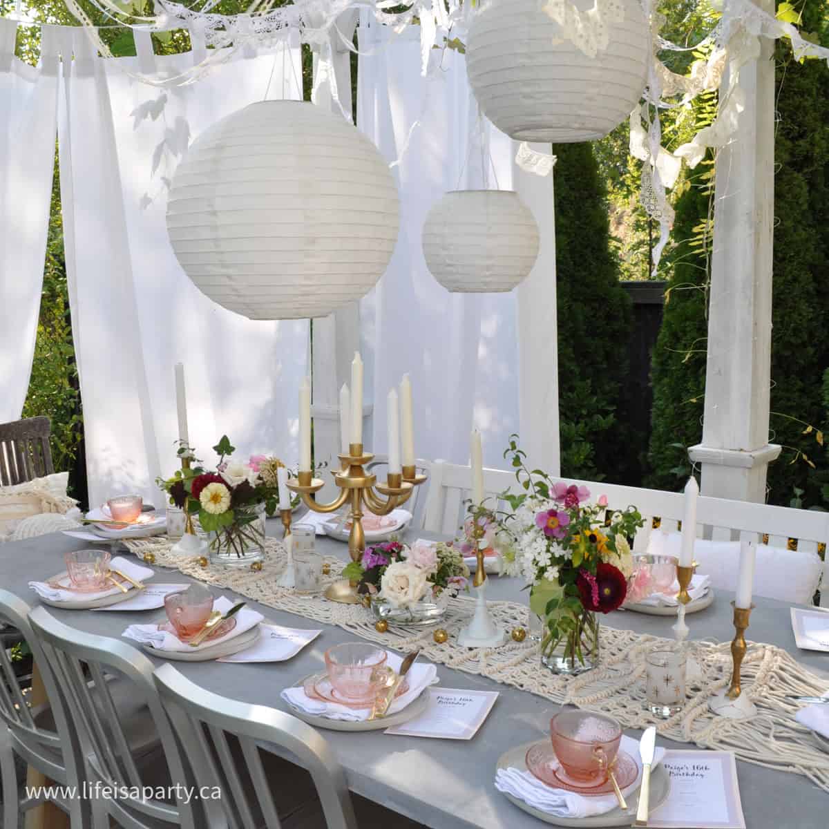 Tea party set up under an outside pergola, with place settings and flowers.