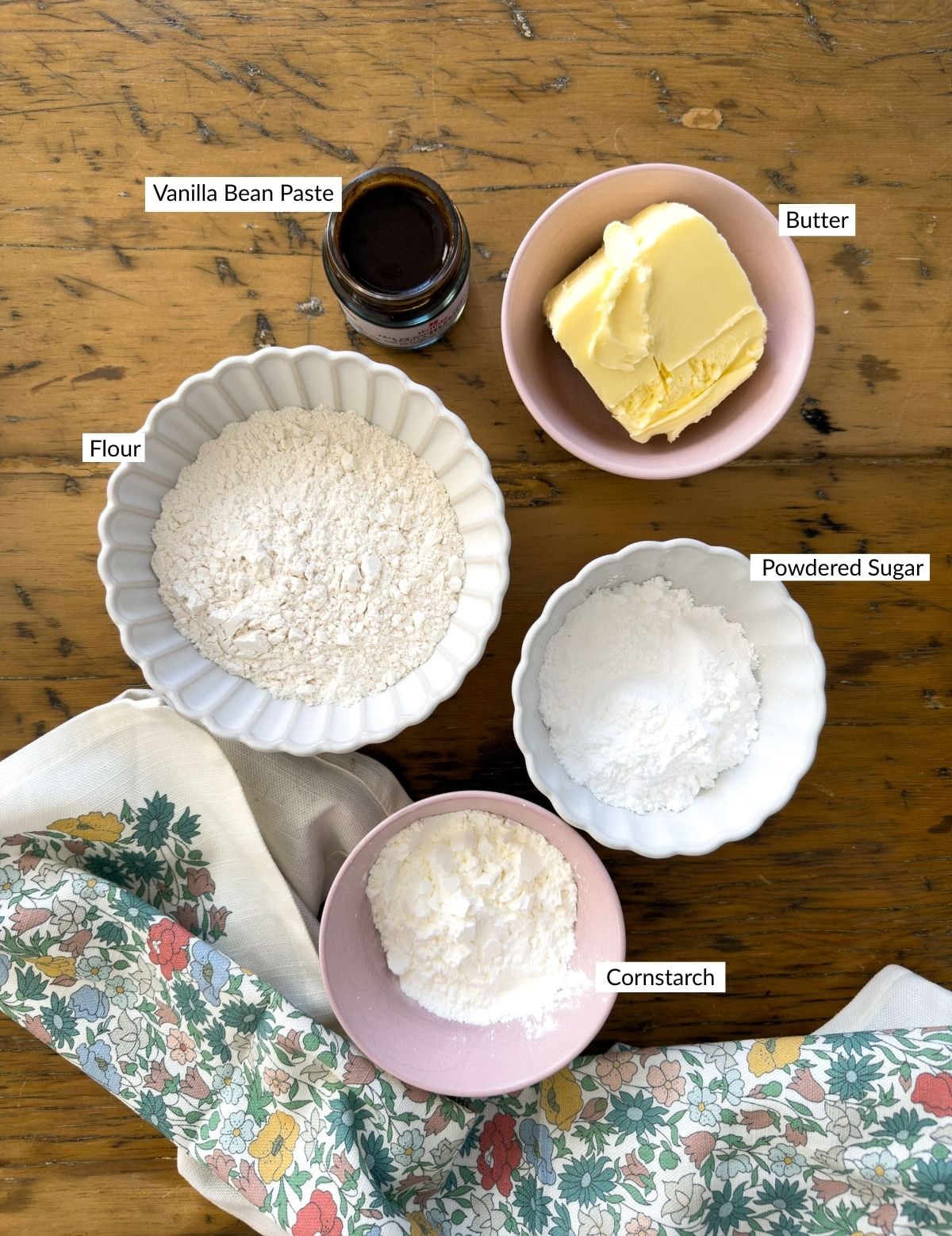 Shortbread cookie ingredients in bowls on a wood table including cornstarch, powdered sugar, flour, butter and vanilla bean paste.