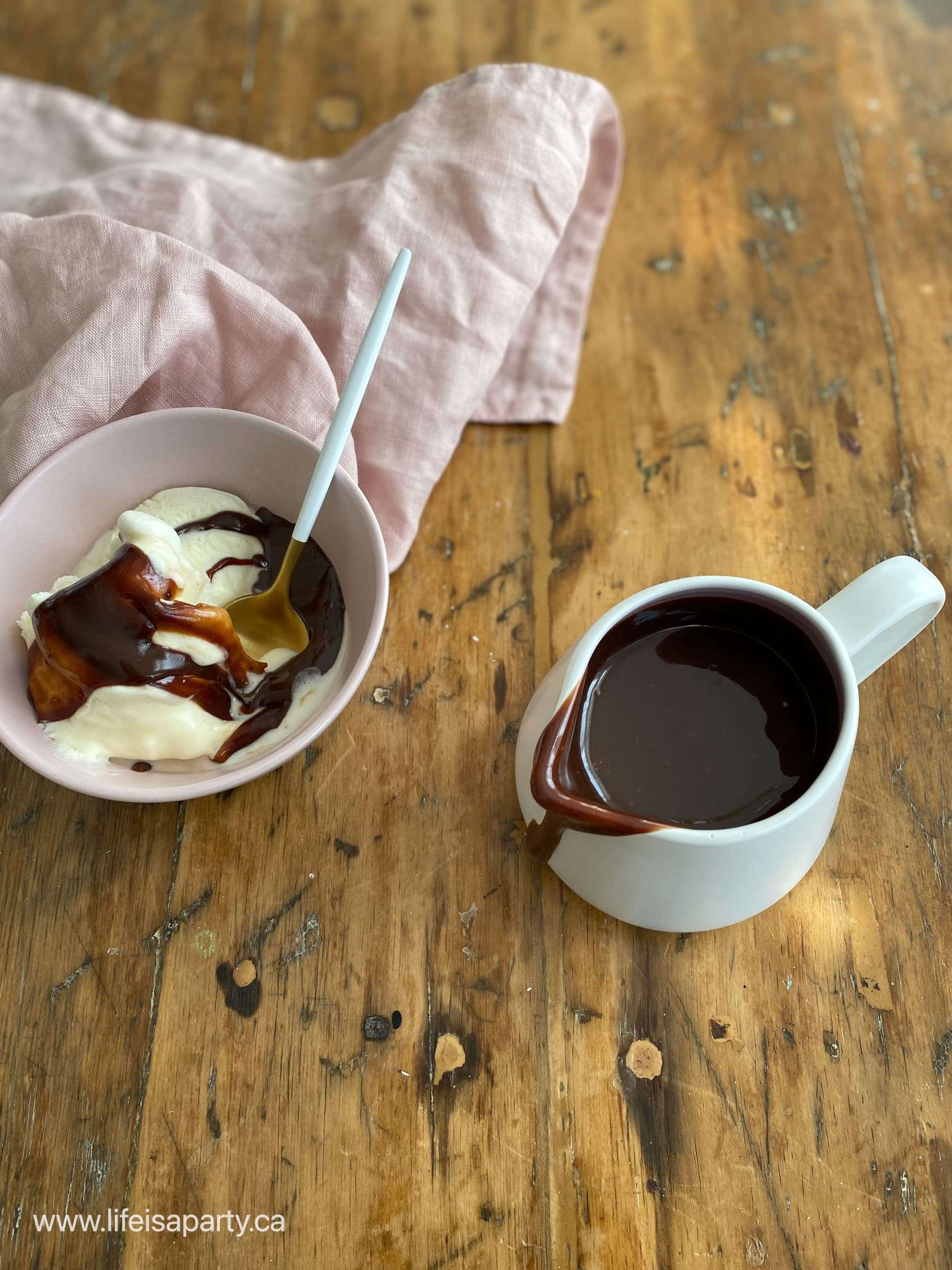 Bowl of vanilla ice cream with hot fudge sauce beside a white pitcher of sauce.