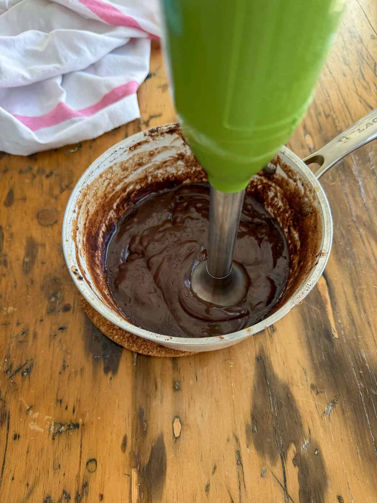 Green immersion blender blending hot fudge sauce directly in a small saucepan on a wood table.