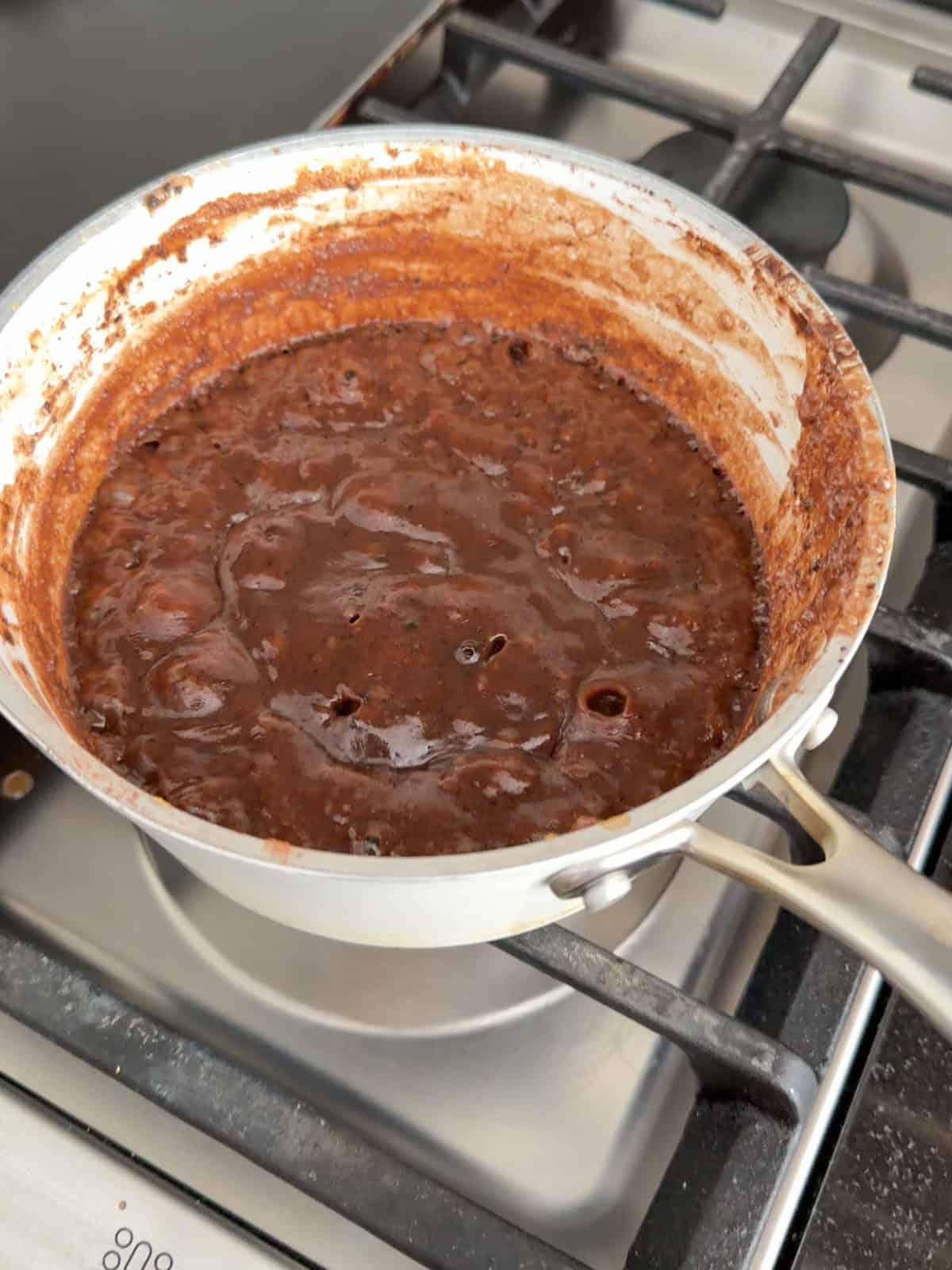 Hot fudge sauce boiling vigorously in a white saucepan on a gas stovetop.