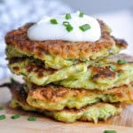 Homemade zucchini pancakes stacked on a wood cutting board, topped with sour cream and chives.