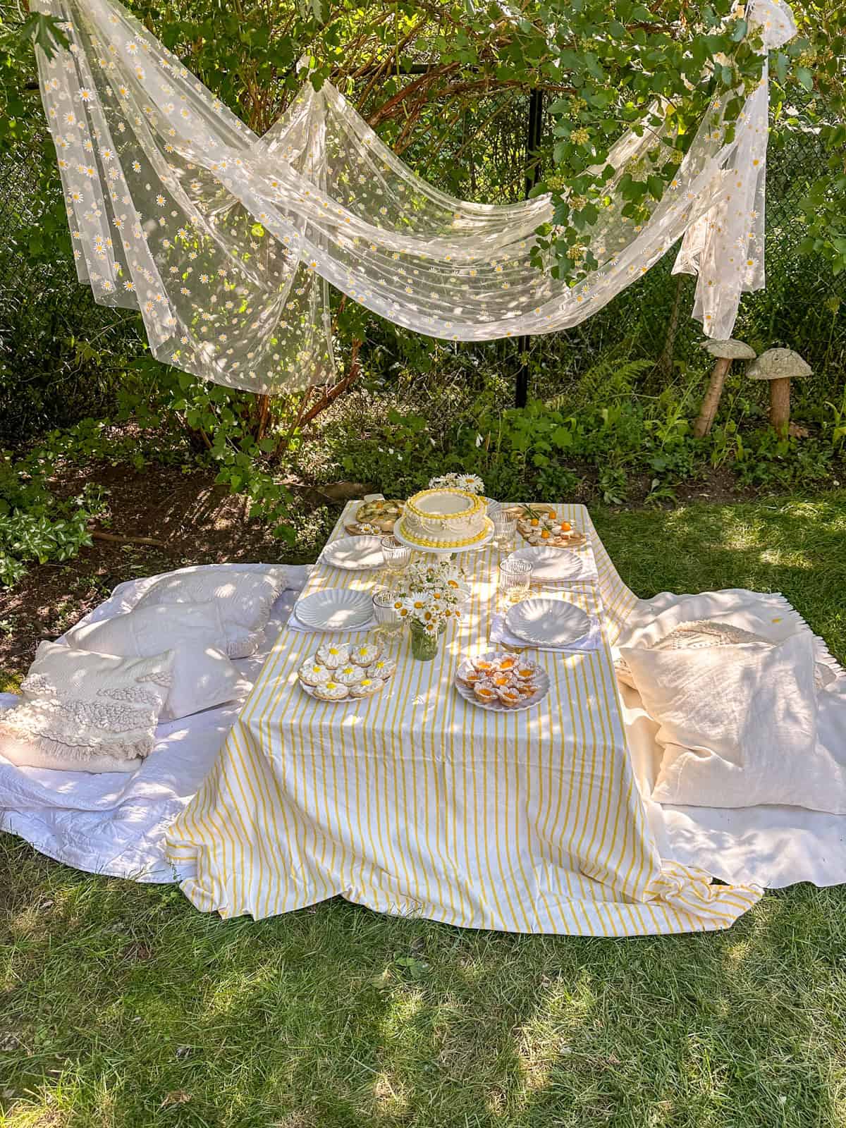 Daisy themed picnic with tulle canopy, and low table with cushions, yellow and white decor, and daisy themed food.