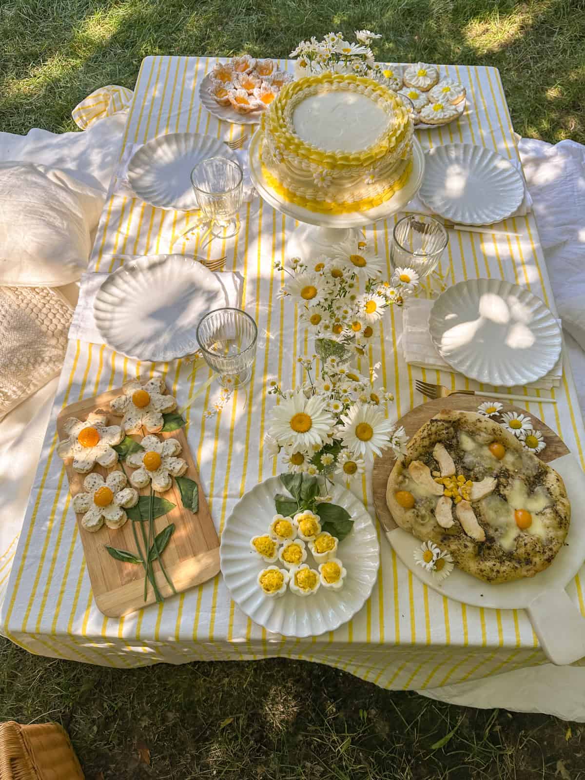 Daisy themed summer picnic party table with yellow and white striped tablecloth, daisy cake, daisy deviled eggs, floral pizza, and fresh daisy flower arrangements.