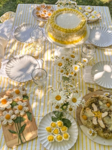 Daisy themed summer picnic party table with yellow and white striped tablecloth, daisy cake, daisy deviled eggs, floral pizza, and fresh daisy flower arrangements.