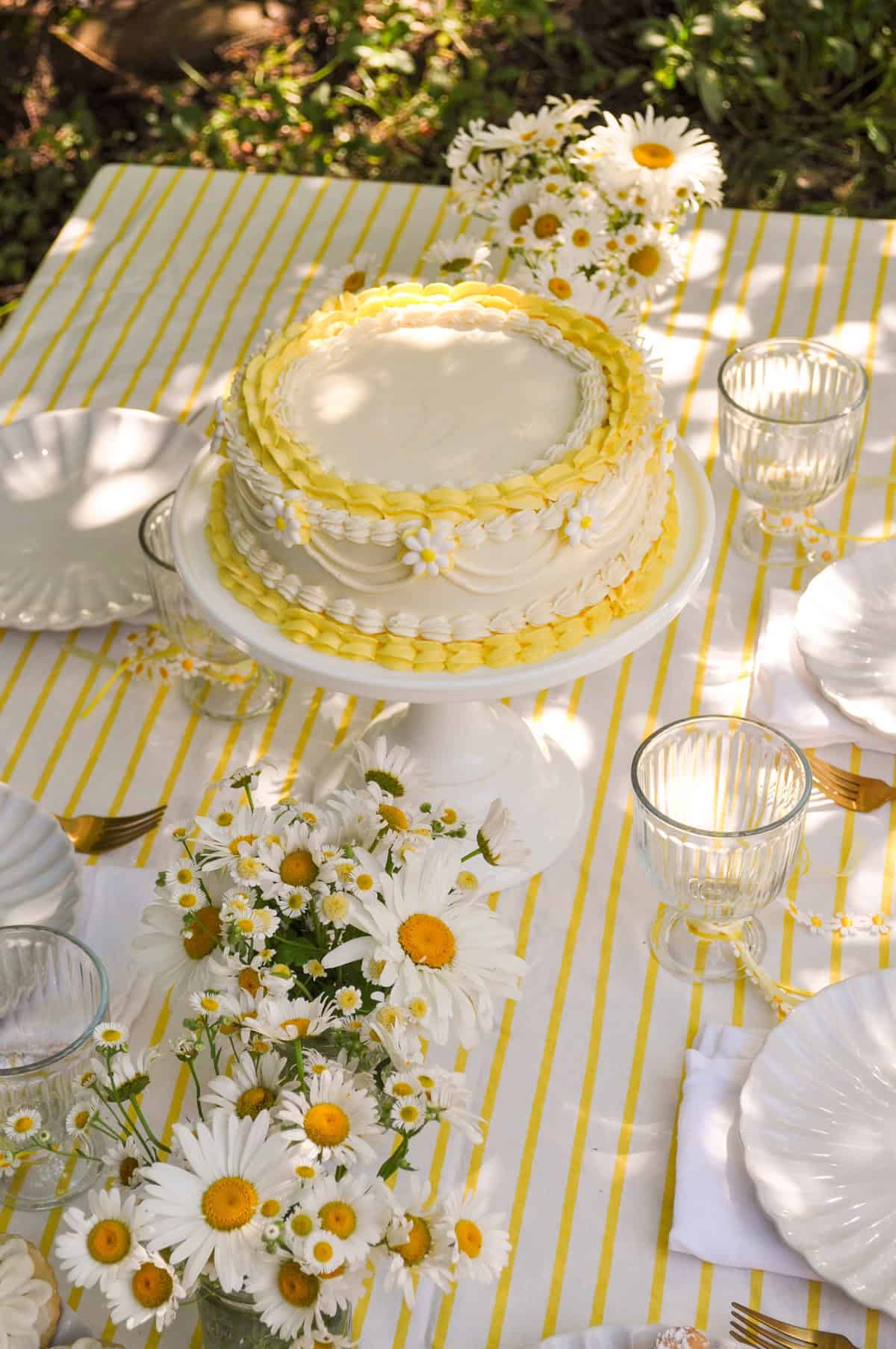 Daisy themed party table with yellow and white striped tablecloth, daisy flower arrangements, and vintage style daisy cake.