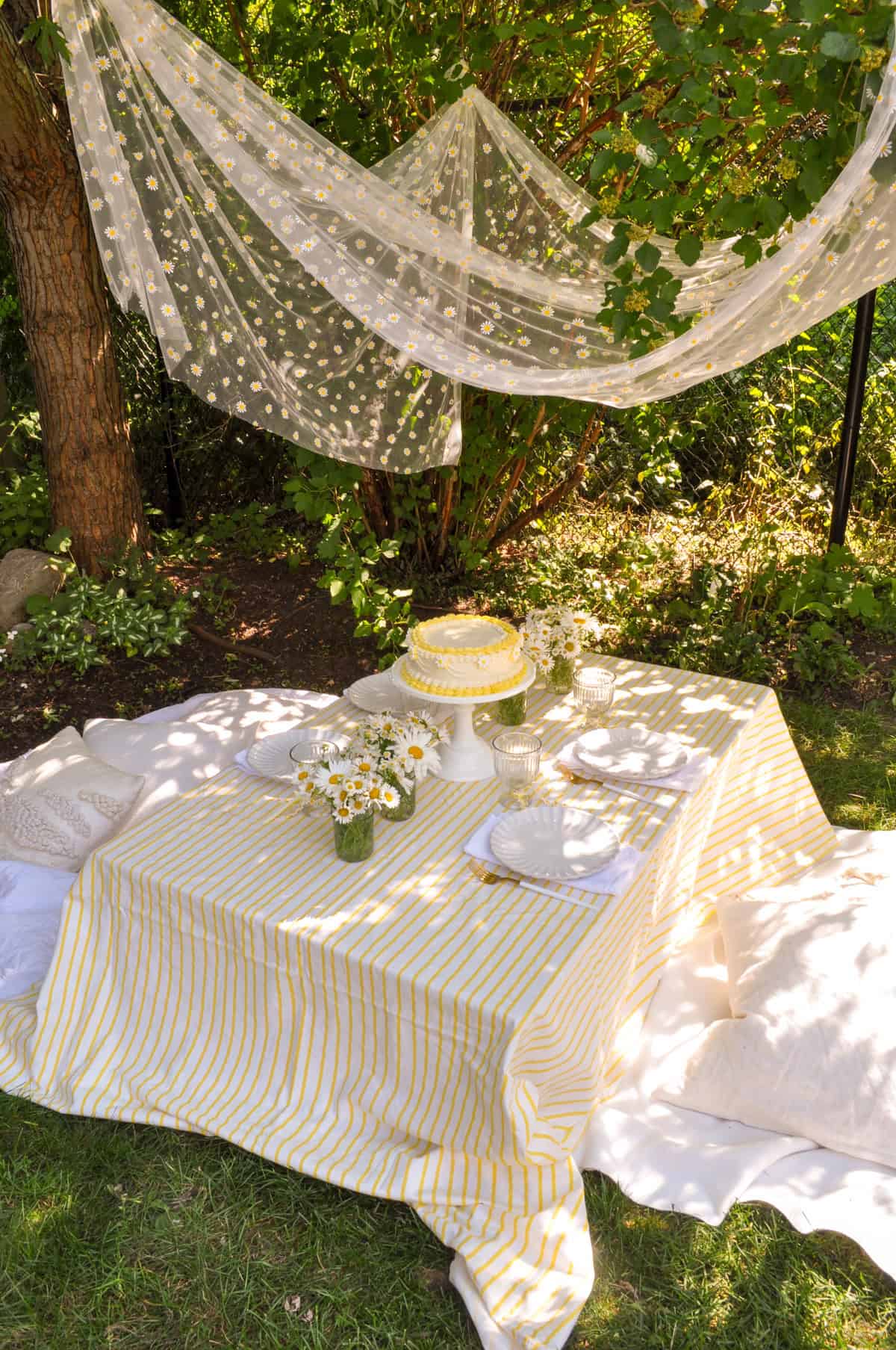 Daisy party table with yellow and white striped tablecloth, daisy flower arrangements, and daisy cake.