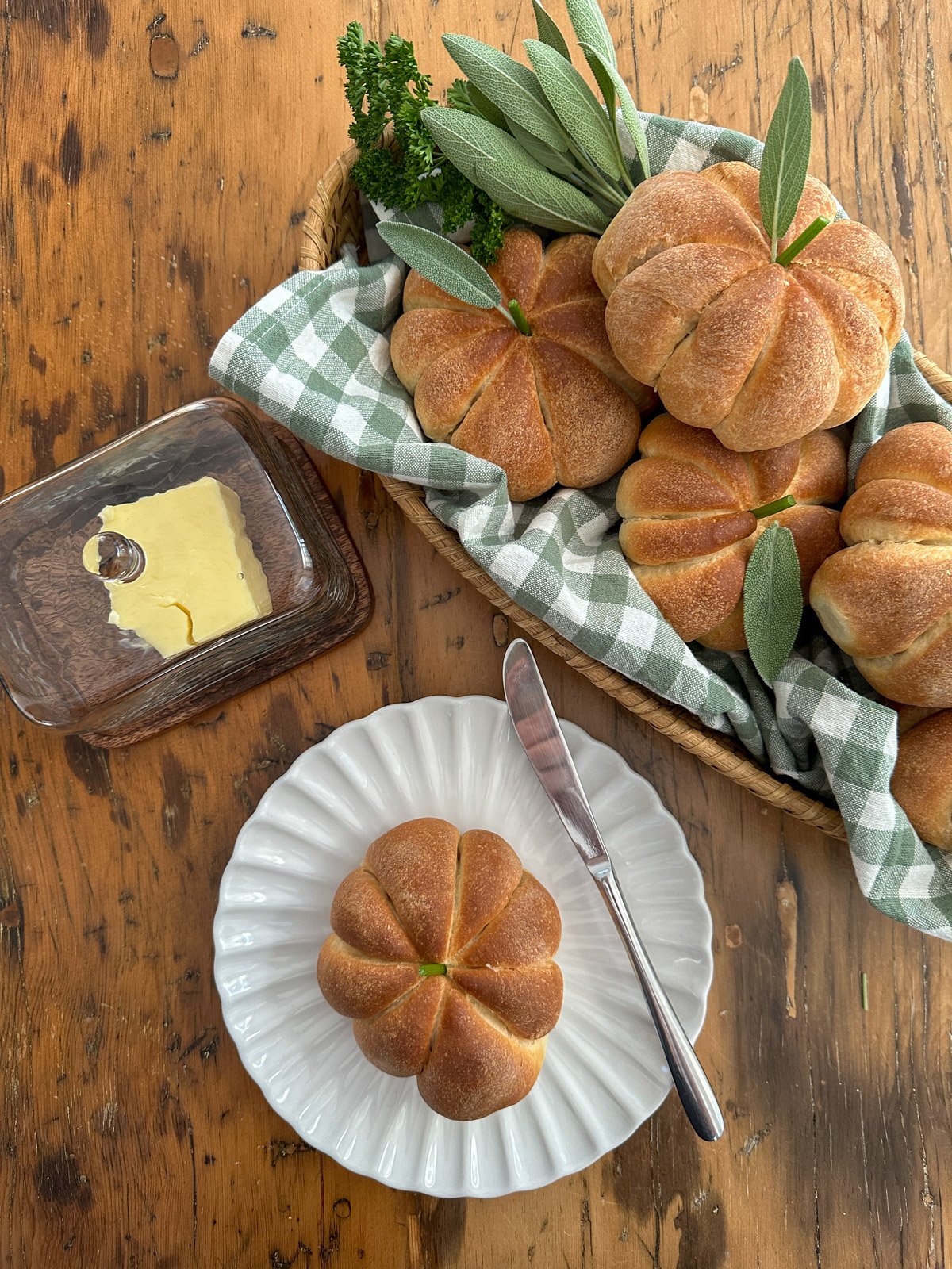 Pumpkin shaped dinner roll with herb stem on plate beside a basket of more rolls.