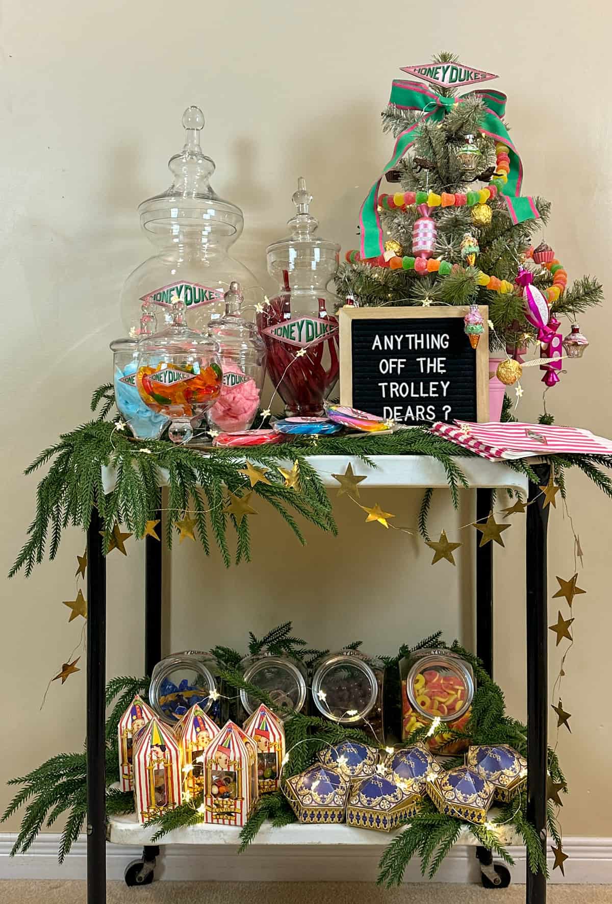 DIY Honeydukes candy cart decorated with garland, candy jars, chocolate frog boxes, and mini tree for Christmas.