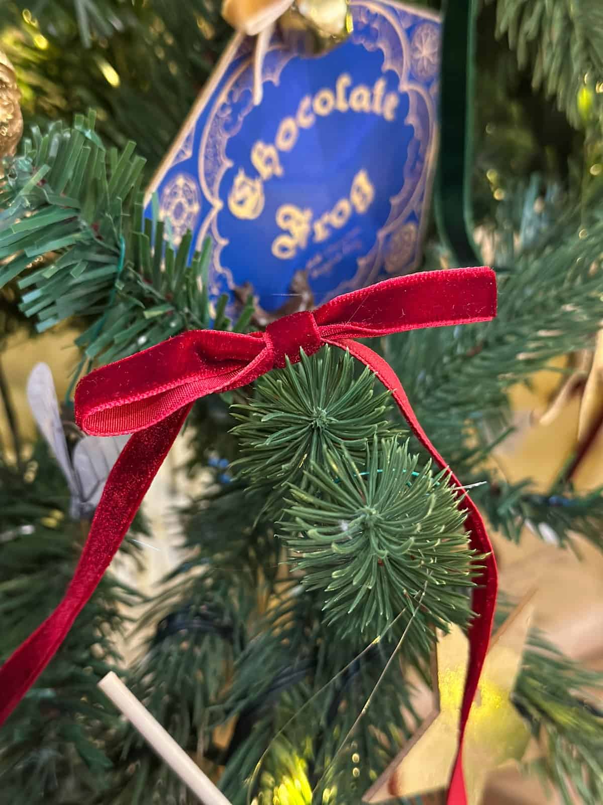 Gryffindor burgundy velvet ribbon bow tied on Harry Potter Christmas tree branch with chocolate frog box ornament in background.