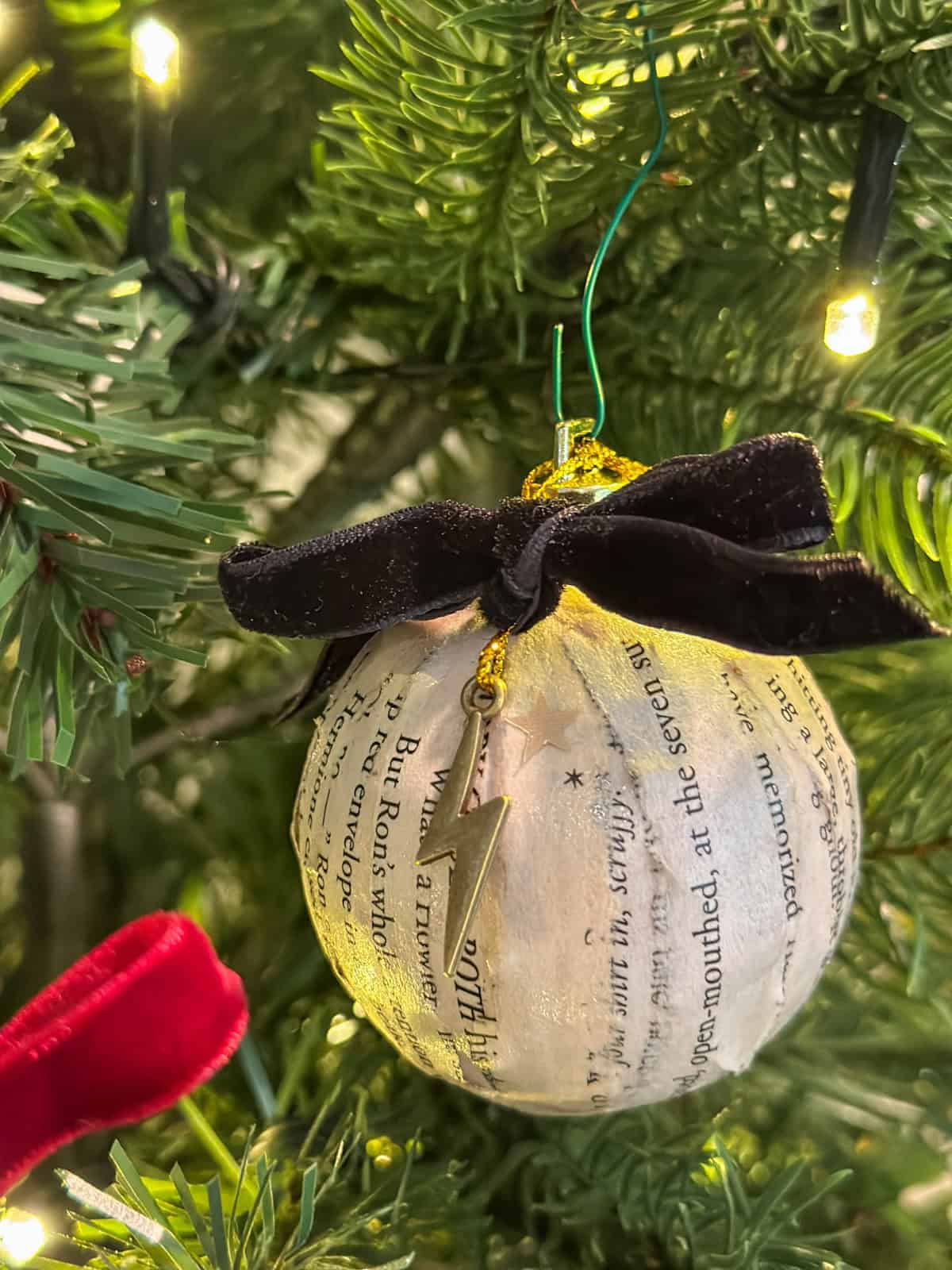 DIY Harry Potter book page Christmas ornament with black velvet bow and gold lightning bolt charm hanging on tree with twinkling lights.