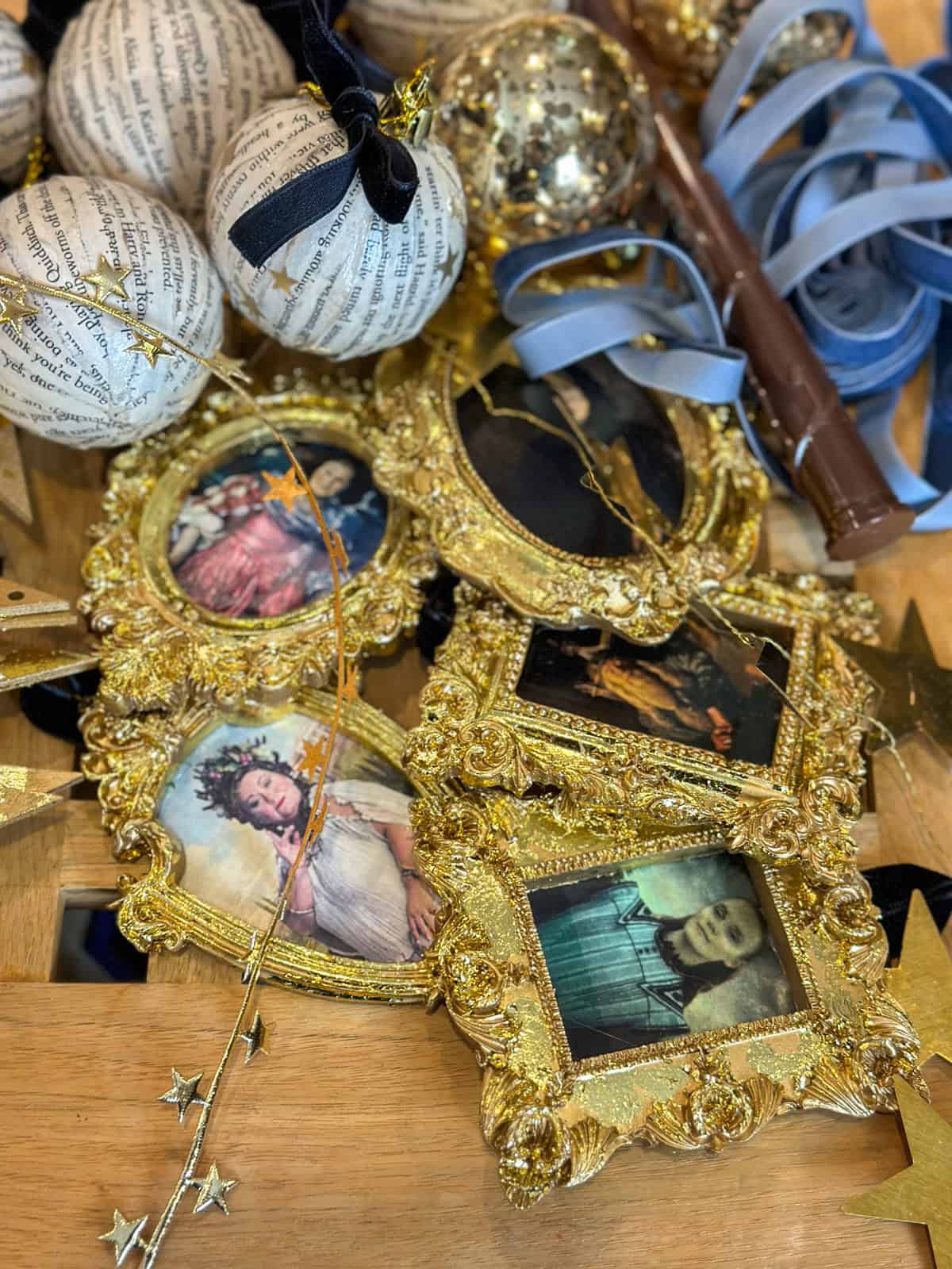Homemade Harry Potter portrait ornaments in ornate gold frames with character images, book page ornaments, house color ribbons, and gold stars on table.