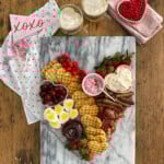 Valentine breakfast board with heart-shaped waffles, heart-shaped eggs, bacon, sausage, fresh berries, and toppings on marble platter.