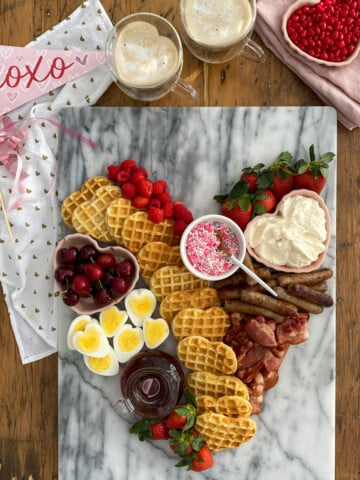 Valentine breakfast board with heart-shaped waffles, heart-shaped eggs, bacon, sausage, fresh berries, and toppings on marble platter.