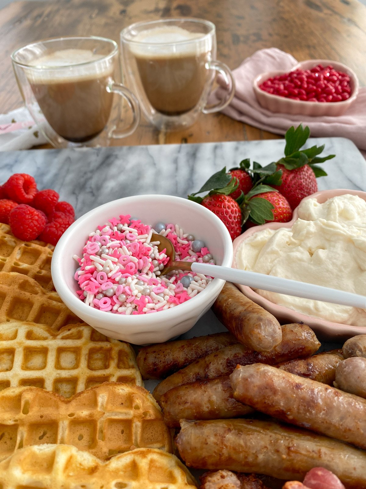 Valentine sprinkles in white bowl with waffles, sausage, fresh berries, and whipped cream for breakfast board.