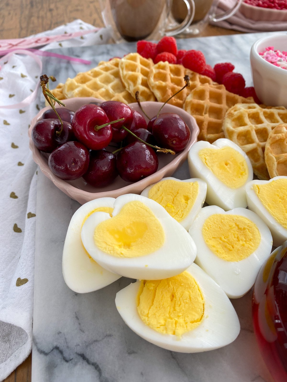 Heart-shaped hard boiled eggs with cherries, heart waffles, and raspberries on Valentine breakfast board.