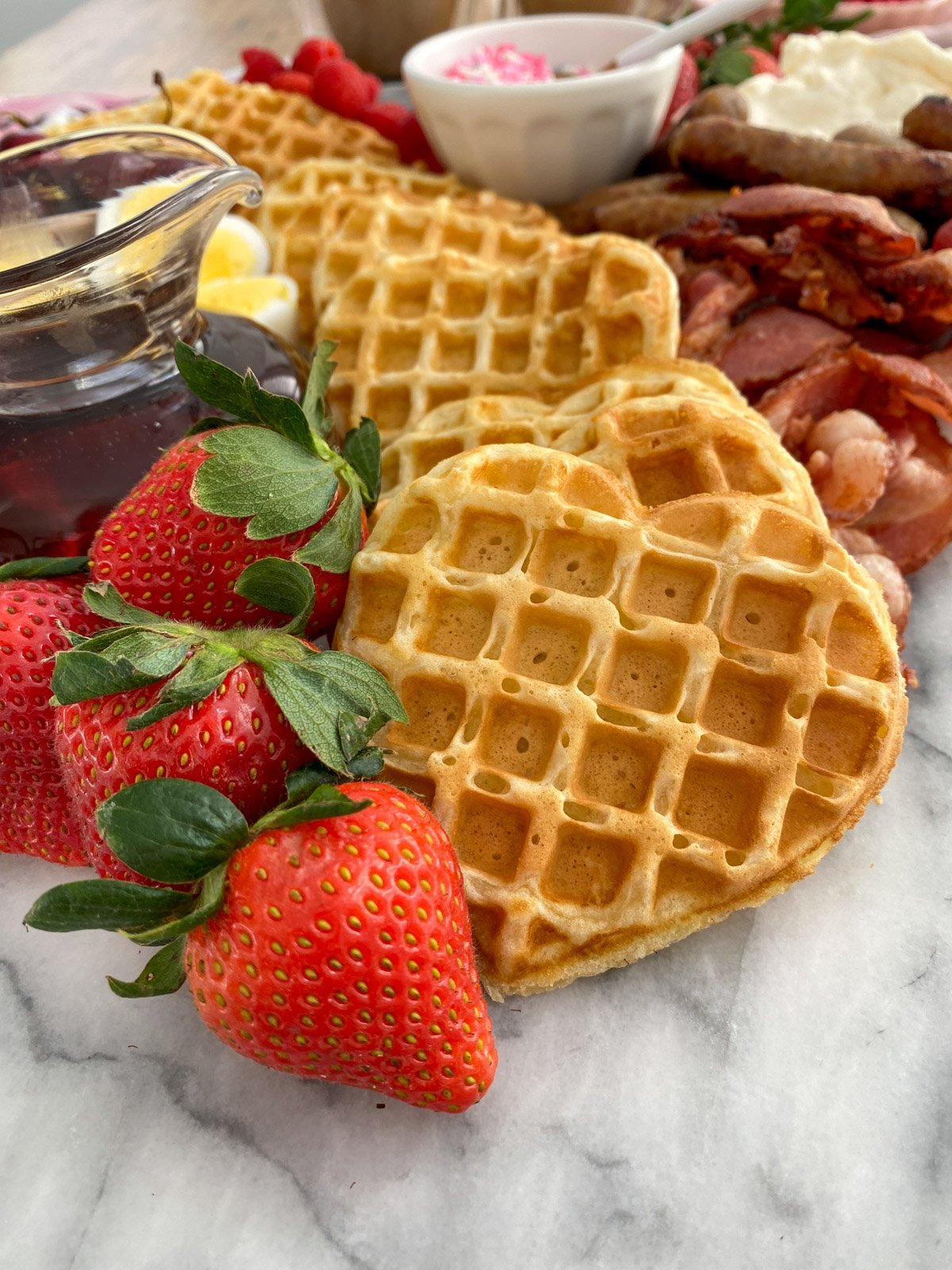 Heart-shaped waffles with fresh strawberries, bacon, sausage, and maple syrup on Valentine breakfast board.