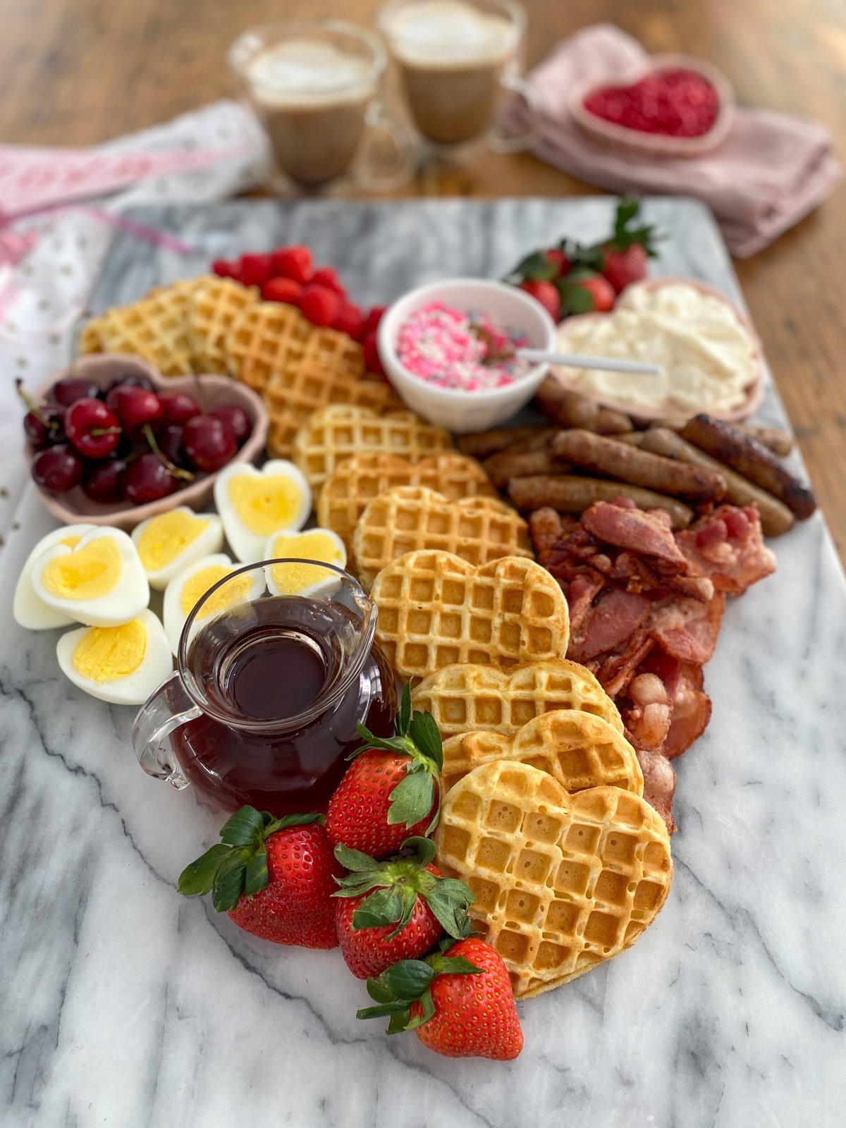 alentine breakfast board arranged in heart shape with waffles, eggs, bacon, sausage, maple syrup, and fresh berries.