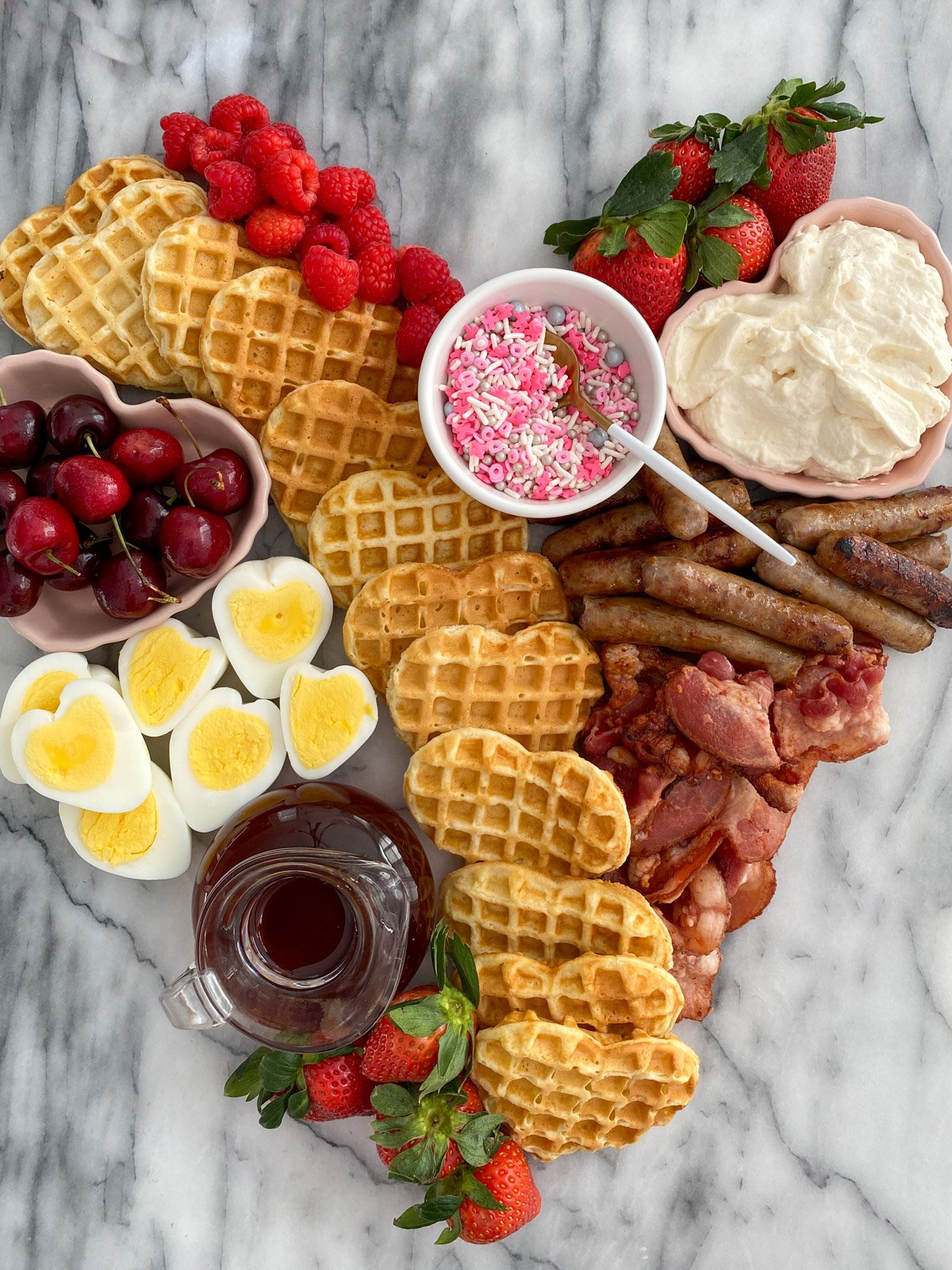 Valentine breakfast board with heart-shaped waffles, heart-shaped eggs, bacon, sausage, fresh berries, and toppings on marble platter.
