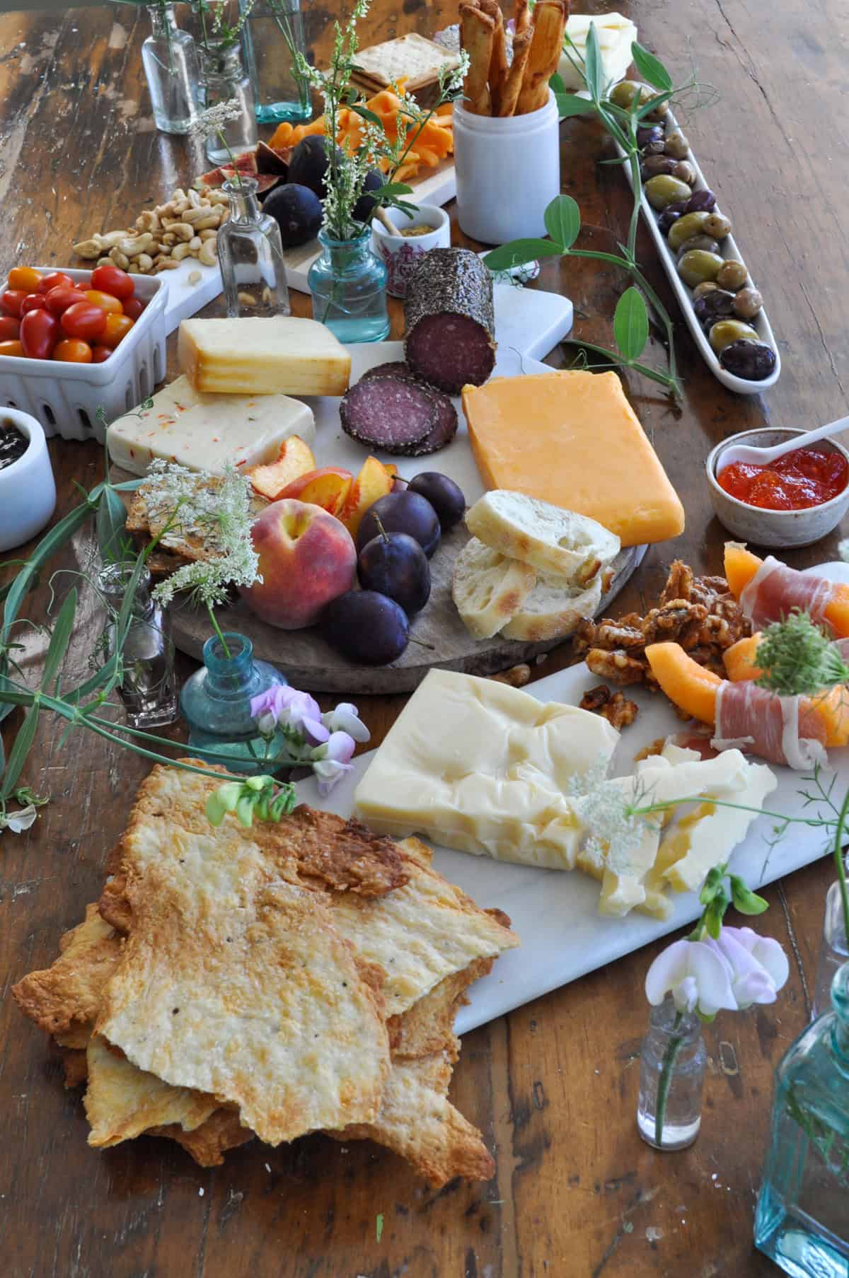 Wine and cheese tasting board on wooden table with various cheeses, crackers, salami, olives, fresh fruit, and small vases with flowers.