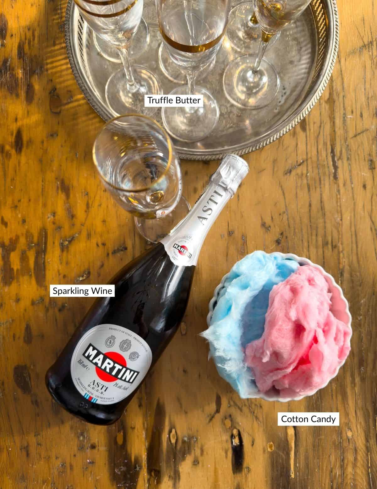 Martini Asti sparkling wine bottle with a bowl of pink and blue cotton candy and gold-rimmed champagne flutes on a wooden table.
