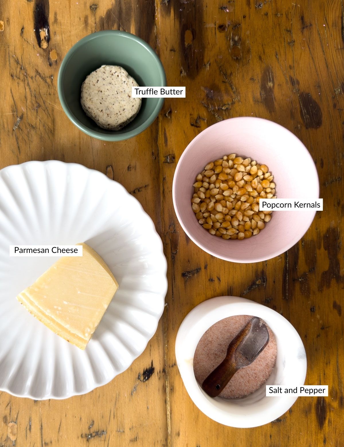 Truffle butter popcorn ingredients arranged on a rustic wood surface including a ball of truffle butter in a green bowl, popcorn kernels in a pink bowl, a wedge of Parmesan on a white plate, and sea salt in a small dish.