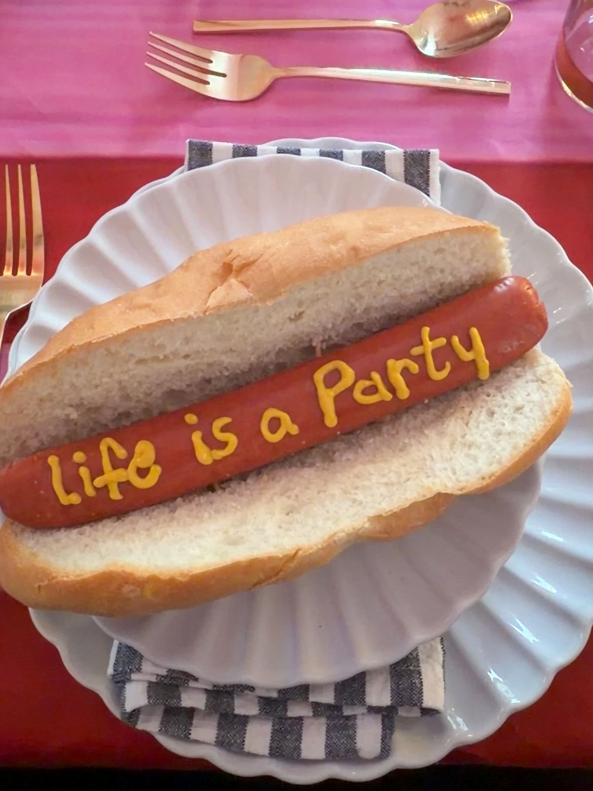 Jumbo beef hot dog in a fresh bakery bun with "Life is a Party" written in mustard, served on a white scalloped plate at a circus themed dinner party.
