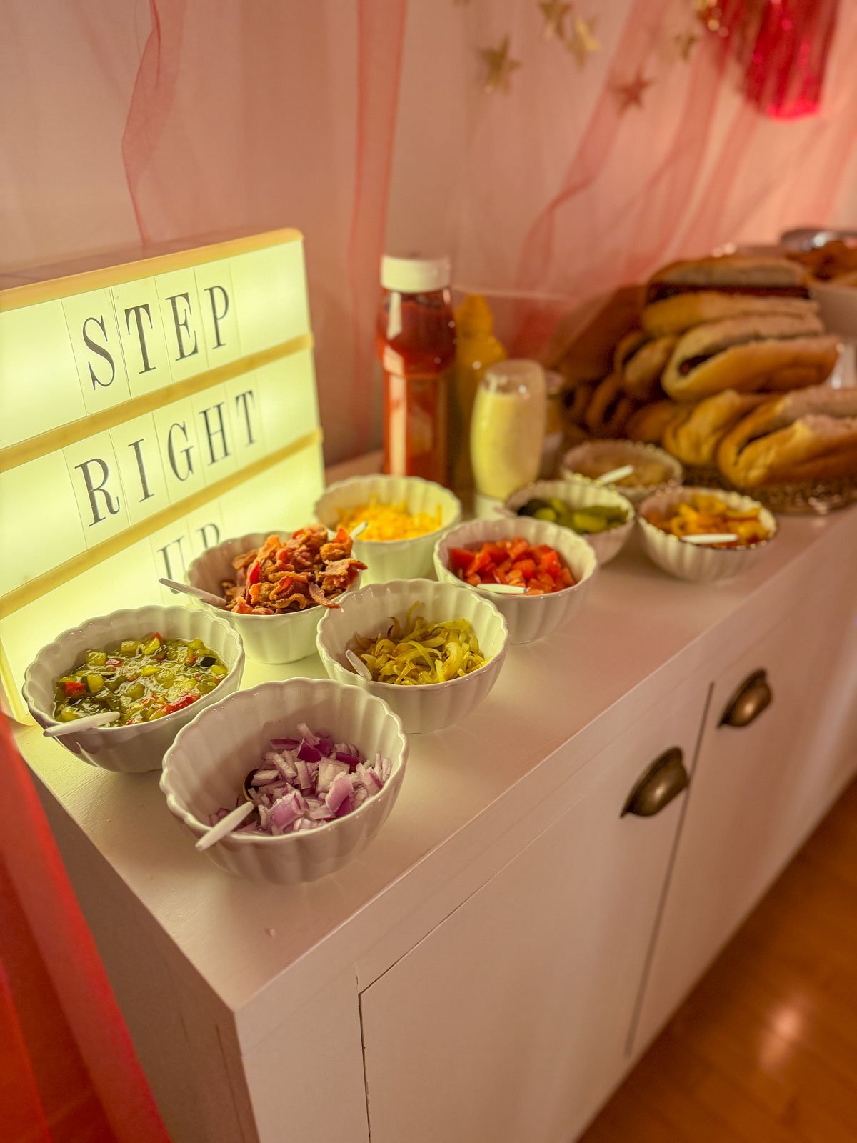Circus party gourmet hot dog buffet toppings including bacon, cheese, relish, tomatoes, pickles, and red onion in white scalloped bowls alongside a "Step Right Up" lightbox sign.